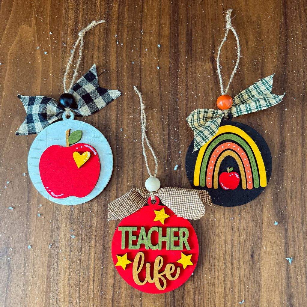 Three handmade wooden teacher-themed hanging ornaments displayed on a brown wood background. One ornament is light blue with a red apple and a black-and-white plaid bow.