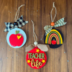 Three handmade wooden teacher-themed hanging ornaments displayed on a brown wood background. One ornament is light blue with a red apple and a black-and-white plaid bow.