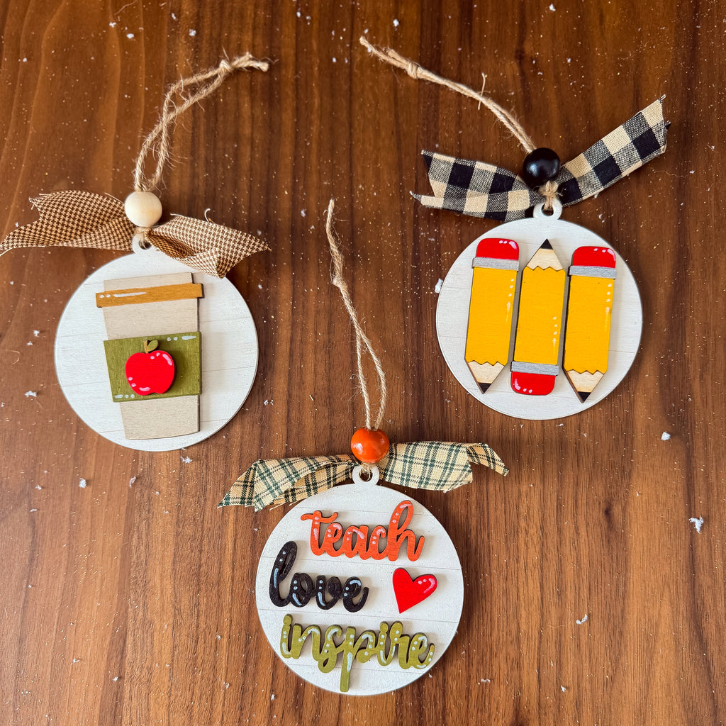 Three round wooden Christmas ornaments with school and teaching themes, displayed on a wooden surface. One ornament features a coffee cup with an apple, another has three yellow pencils, and the third says 'teach love inspire' in decorative lettering with a plaid ribbon.