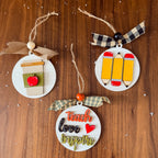 Three round wooden Christmas ornaments with school and teaching themes, displayed on a wooden surface. One ornament features a coffee cup with an apple, another has three yellow pencils, and the third says 'teach love inspire' in decorative lettering with a plaid ribbon.