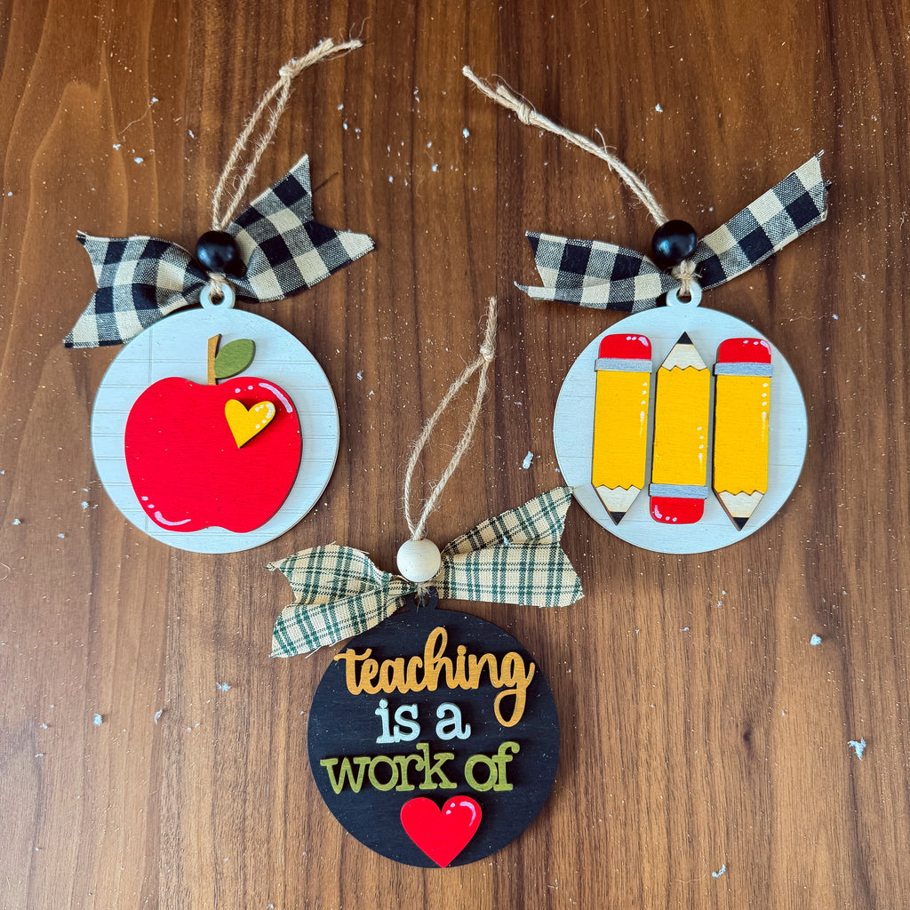 Three circular wooden teacher-themed ornaments hanging on a brown wood surface. One ornament shows a red apple with a small yellow heart and a black-and-white plaid ribbon bow.
