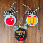 Three circular wooden teacher-themed ornaments hanging on a brown wood surface. One ornament shows a red apple with a small yellow heart and a black-and-white plaid ribbon bow.