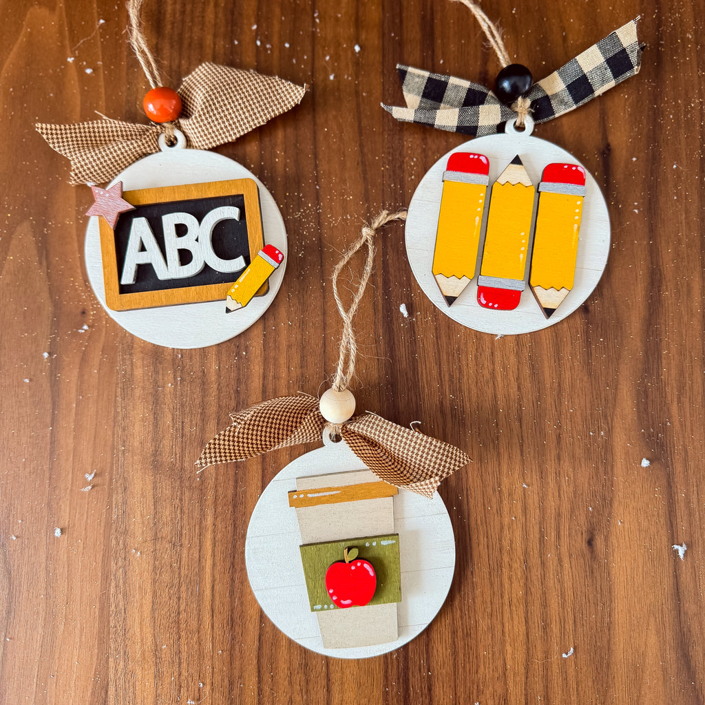 Three round wooden school-themed ornaments on a brown wood background. One ornament has a black chalkboard with 'ABC' and a pencil, decorated with a beige burlap ribbon.