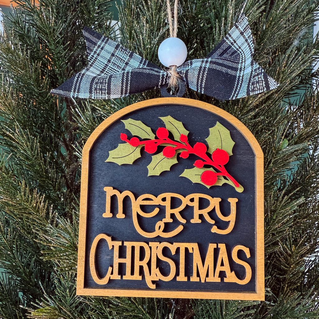 A Christmas ornament shaped like a wooden tombstone hangs on a pine tree. The ornament has a dark charcoal background with raised gold lettering that reads “merry CHRISTMAS,” a green and red holly branch across the top, and a black-and-white plaid ribbon bow with a white bead at the hanging loop.