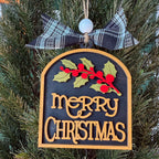 A Christmas ornament shaped like a wooden tombstone hangs on a pine tree. The ornament has a dark charcoal background with raised gold lettering that reads “merry CHRISTMAS,” a green and red holly branch across the top, and a black-and-white plaid ribbon bow with a white bead at the hanging loop.