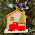 A wooden house-shaped Christmas ornament hanging on a pine tree, featuring a red pickup truck carrying a green Christmas tree, with a black-and-white buffalo plaid ribbon bow at the top.