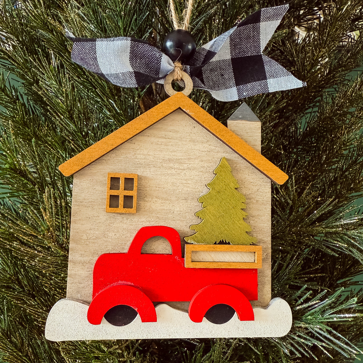 A wooden house-shaped Christmas ornament hanging on a pine tree, featuring a red pickup truck carrying a green Christmas tree, with a black-and-white buffalo plaid ribbon bow at the top.