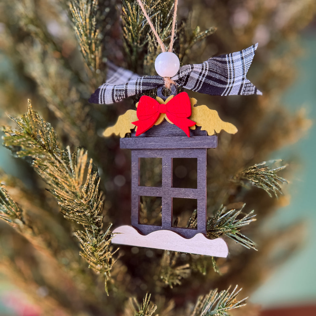A handmade wooden Christmas ornament shaped like a house with a black window frame, a red felt bow, gold felt wings, a plaid ribbon, and a white snow base, hanging on a Christmas tree.