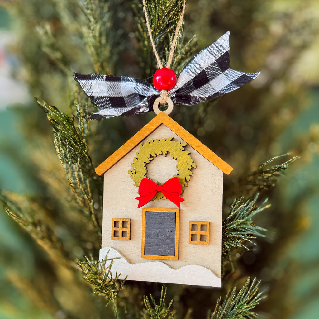 A wooden house-shaped Christmas ornament hanging on a pine tree. The ornament has a beige body, a yellow-orange roof, a green wreath, a red bow, and a black-and-white checkered ribbon with a red bead at the top.