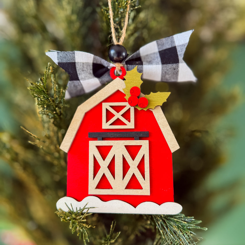 A red barn-shaped Christmas ornament hanging on a green Christmas tree. The ornament has a black-and-white buffalo plaid ribbon bow at the top, a gold holly leaf with red berries, and a white snowy base.