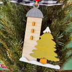 A wooden Christmas ornament shaped like a lighthouse next to a pine tree, hanging on a green Christmas tree branch.