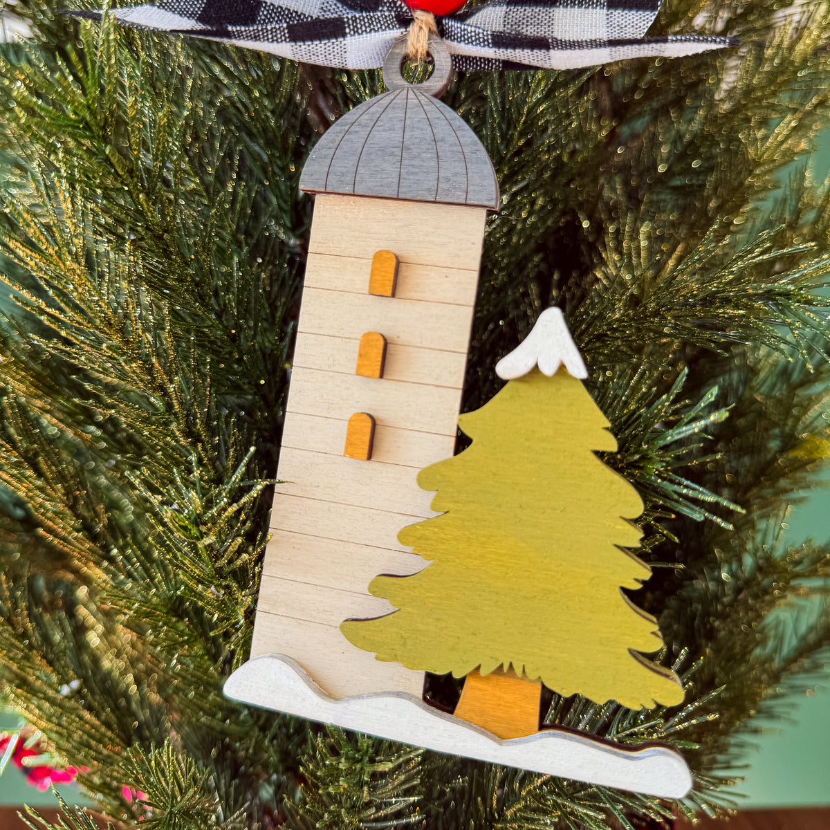 A wooden Christmas ornament shaped like a lighthouse next to a pine tree, hanging on a green Christmas tree branch.