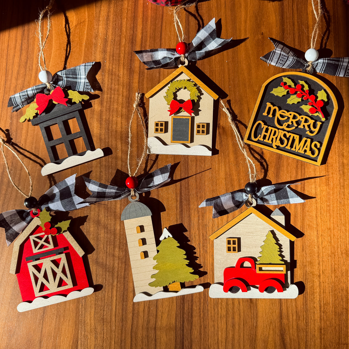 Six wooden Christmas house ornaments with plaid ribbon hangers are arranged on a warm wood surface. The ornaments feature different holiday designs including a barn, a lighthouse, a house with a Christmas tree, a house with a red truck, a wreath, and a 'Merry Christmas' sign.