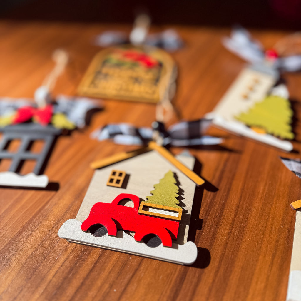 A close-up of a wooden Christmas ornament shaped like a house with a red pickup truck carrying a green Christmas tree, hanging on a wooden surface with other ornaments blurred in the background.