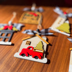 A close-up of a wooden Christmas ornament shaped like a house with a red pickup truck carrying a green Christmas tree, hanging on a wooden surface with other ornaments blurred in the background.