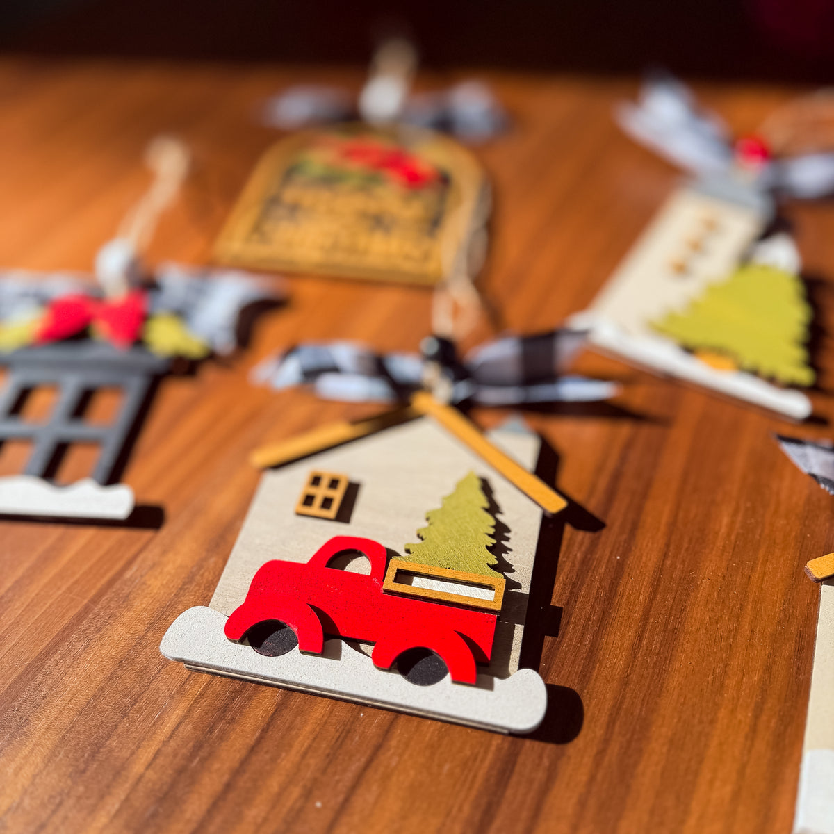 A close-up of a wooden Christmas ornament shaped like a house with a red pickup truck carrying a green Christmas tree, hanging on a wooden surface with other ornaments blurred in the background.