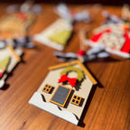 A close-up of a small house-shaped Christmas ornament on a wooden surface, with other blurred ornaments in the background. The ornament is beige with a dark gray door, a green wreath, a red bow, and small brown window details.