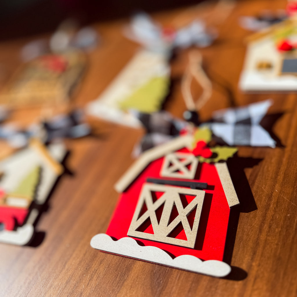 A close-up of a red barn-shaped ornament on a wooden surface, with other decorative ornaments blurred in the background.