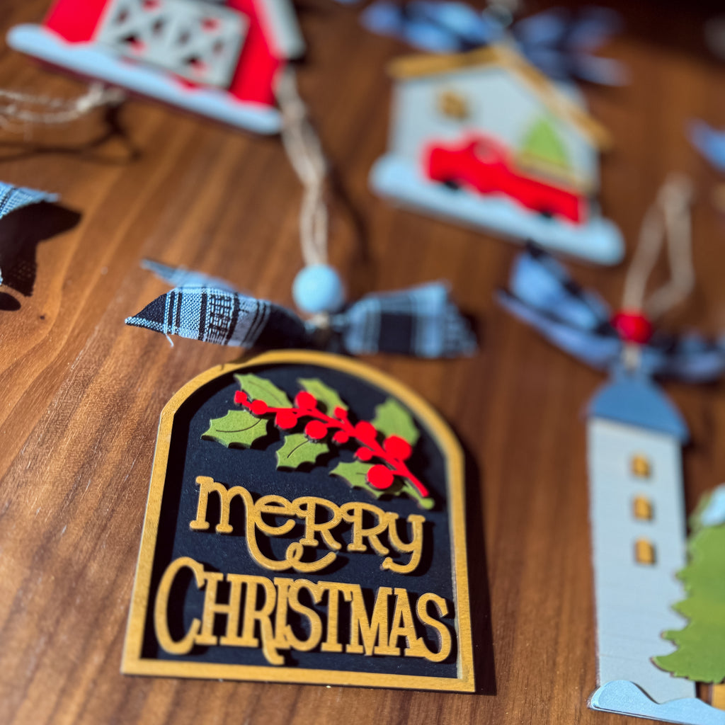 A close-up of a wooden Christmas ornament on a wooden tabletop. The ornament is arched with a black center panel, gold border, gold 'Merry Christmas' lettering, green holly leaves, and red berries.
