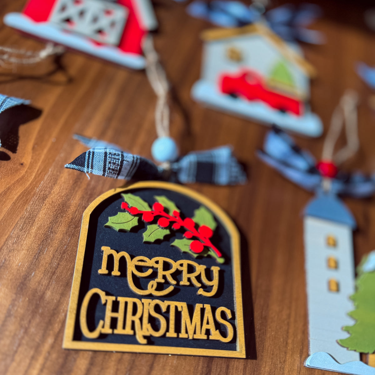 A close-up of a wooden Christmas ornament on a wooden tabletop. The ornament is arched with a black center panel, gold border, gold 'Merry Christmas' lettering, green holly leaves, and red berries.