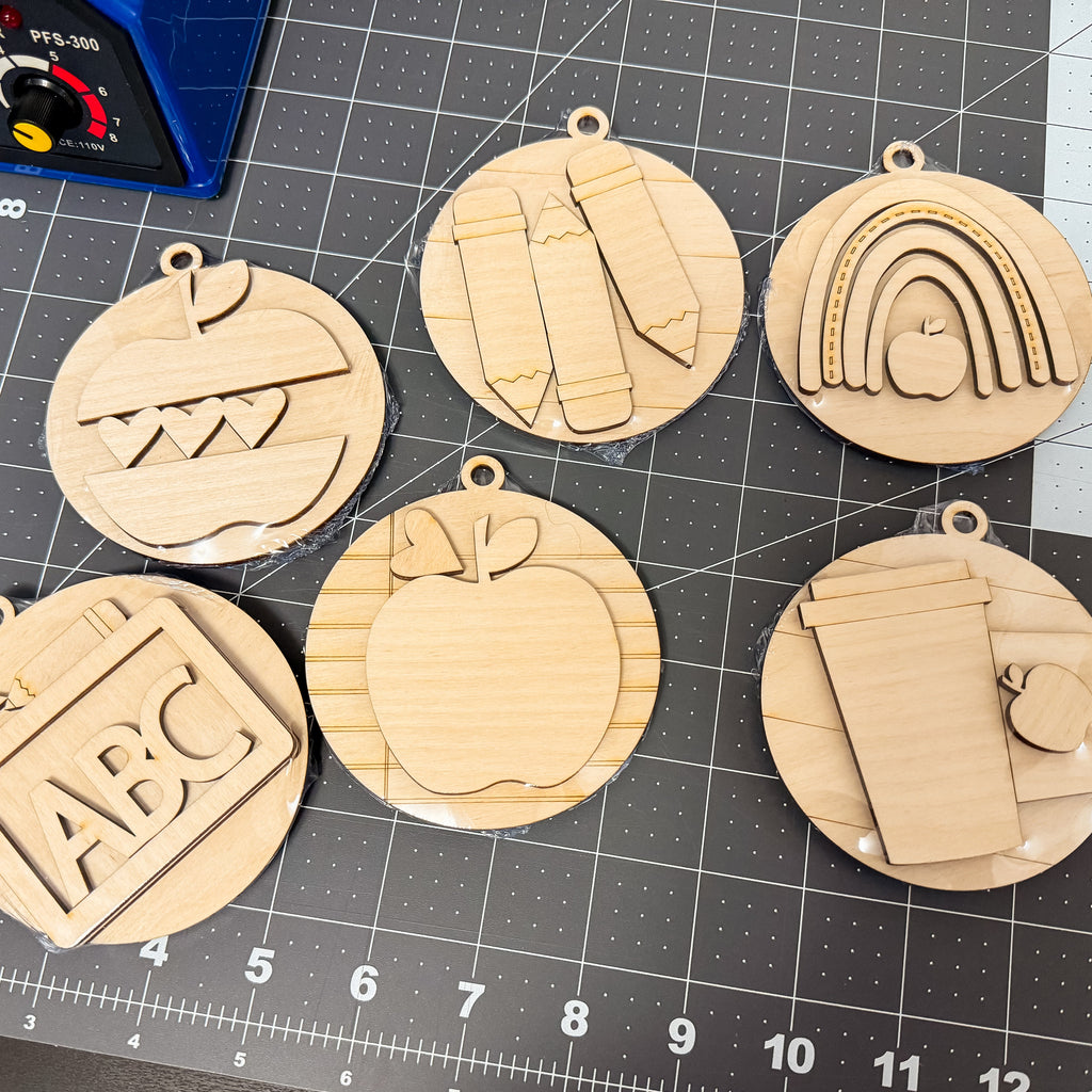 Six round laser-cut wooden ornaments with school-themed designs, including a rainbow, apple, alphabet, and coffee cup, arranged on a black cutting mat.