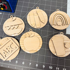Six round laser-cut wooden ornaments with school-themed designs, including a rainbow, apple, alphabet, and coffee cup, arranged on a black cutting mat.