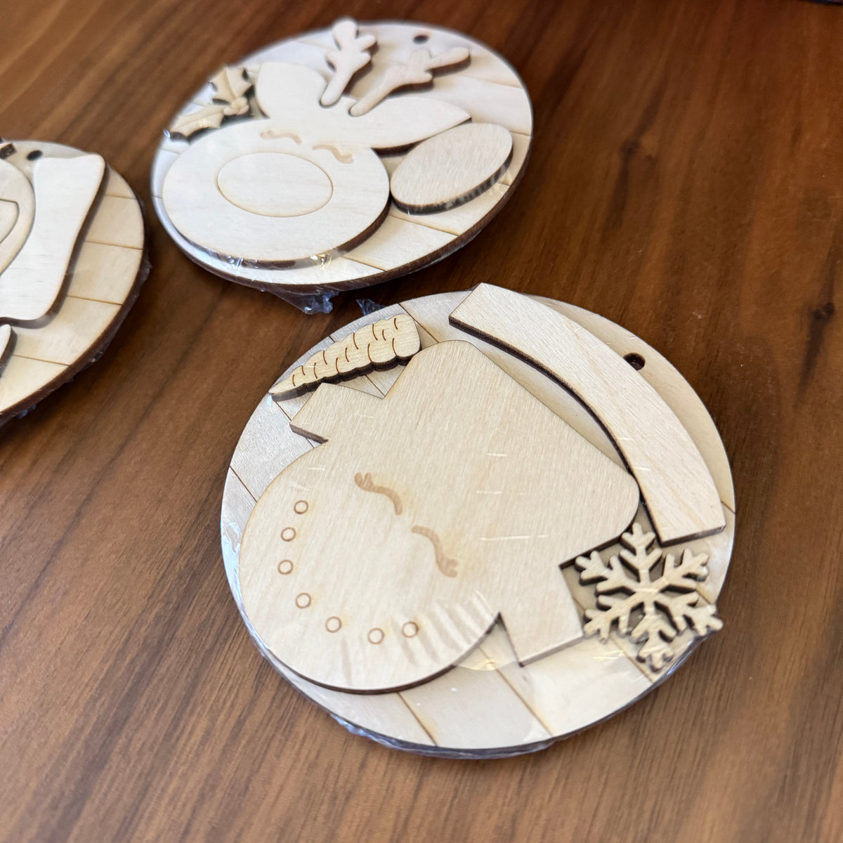 Unpainted laser-cut wooden ornaments or craft pieces arranged on a wooden tabletop, including a snowman face, star, oval shapes, and a snowflake detail.