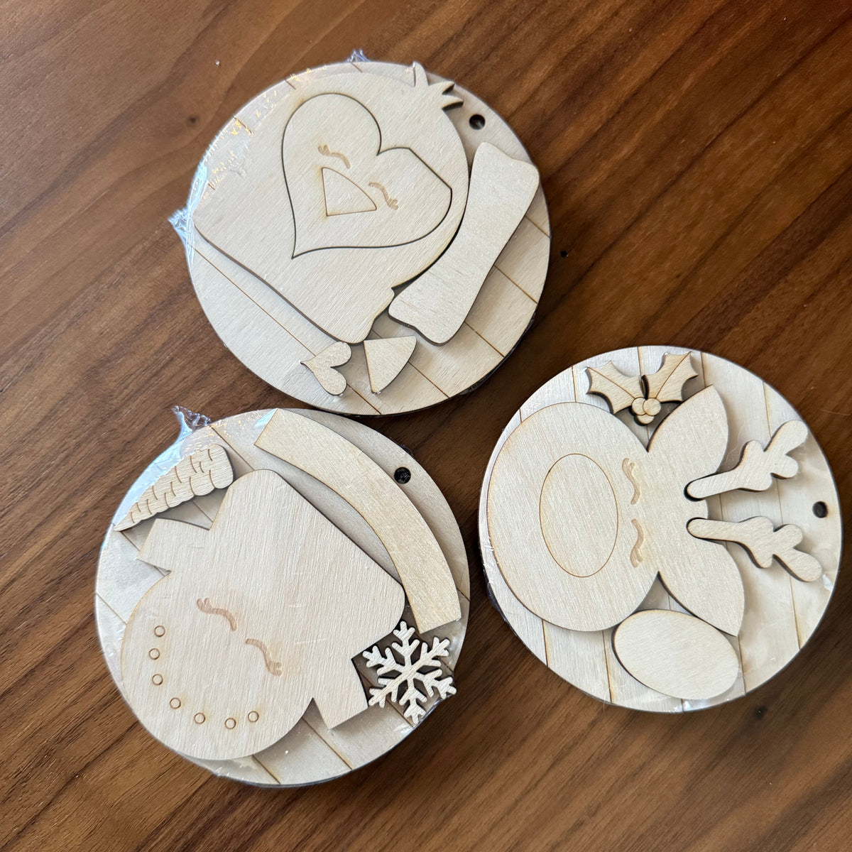 Three round wooden craft ornaments with laser-cut holiday designs, including a heart, snowman, and reindeer, displayed on a wooden tabletop.