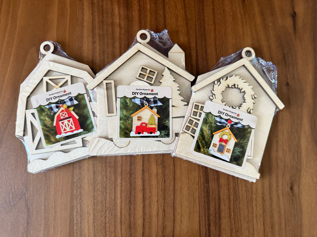 Three unpainted wooden house-shaped DIY ornaments in plastic wrapping, displayed on a wooden tabletop. Each ornament features a different house design with a printed card insert showing a finished example.