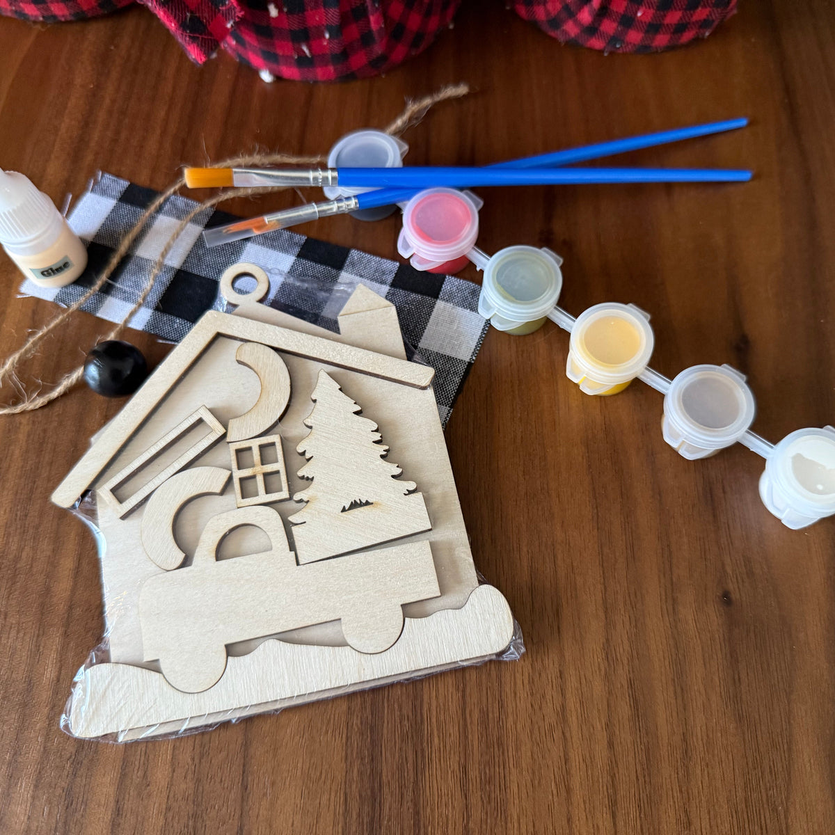 A DIY wooden house craft kit laid out on a wooden table, including laser-cut wooden house pieces, a plaid fabric backdrop, small paint pots, two paintbrushes, and a glue bottle.