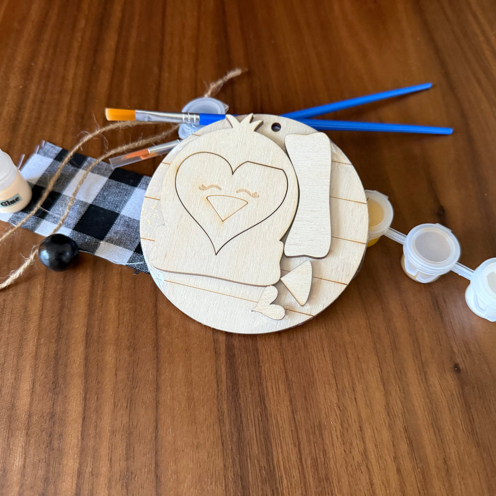 A wooden craft kit featuring a round unfinished wood plaque with a heart-shaped cutout, accompanied by small paint bottles, paintbrushes, twine, and a black-and-white buffalo plaid ribbon, all arranged on a wooden tabletop.