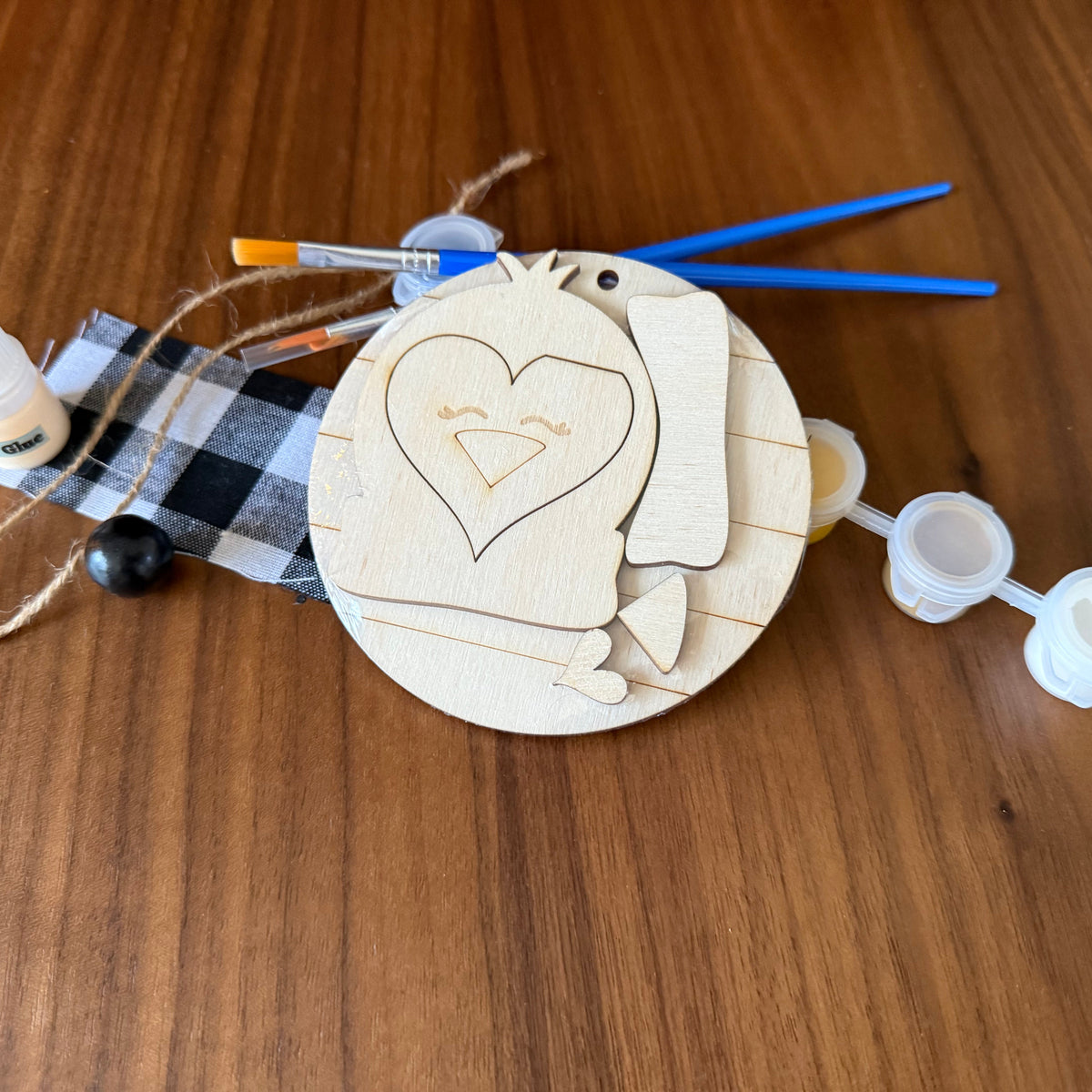 A wooden craft kit featuring a round unfinished wood plaque with a heart-shaped cutout, accompanied by small paint bottles, paintbrushes, twine, and a black-and-white buffalo plaid ribbon, all arranged on a wooden tabletop.