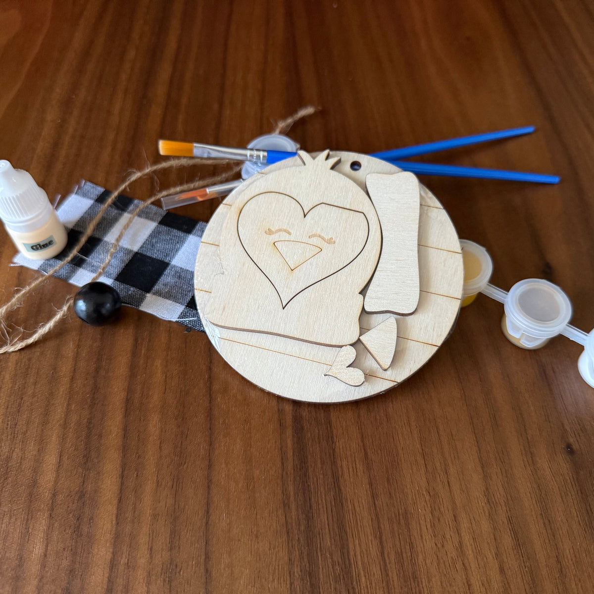 A wooden bird-shaped craft decoration or ornament laid on a brown wood-grain surface, with attached twine, a black-and-white plaid ribbon, a small white bottle, a black bead, two blue-handled tools, and white plastic caps.