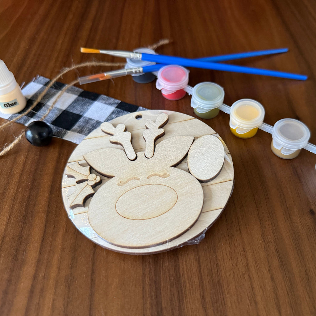 A wooden reindeer craft kit laid out on a wooden table, including laser-cut wooden reindeer pieces on a round base, small paint pots in multiple colors, two blue-handled paintbrushes, a bottle of wood glue, twine, and a black-and-white check cloth.