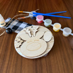 A wooden reindeer craft kit laid out on a wooden table, including laser-cut wooden reindeer pieces on a round base, small paint pots in multiple colors, two blue-handled paintbrushes, a bottle of wood glue, twine, and a black-and-white check cloth.