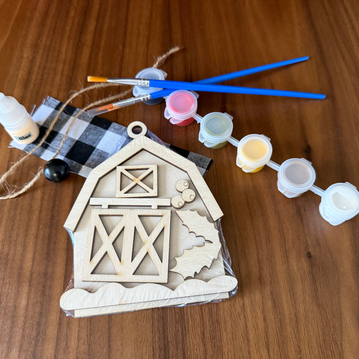 A DIY wooden ornament kit featuring a pre-cut unfinished wooden barn ornament, a small bottle of white paint, a black and white buffalo plaid fabric bag, twine, and four small paint pots with blue-handled paintbrushes, all arranged on a wooden tabletop.