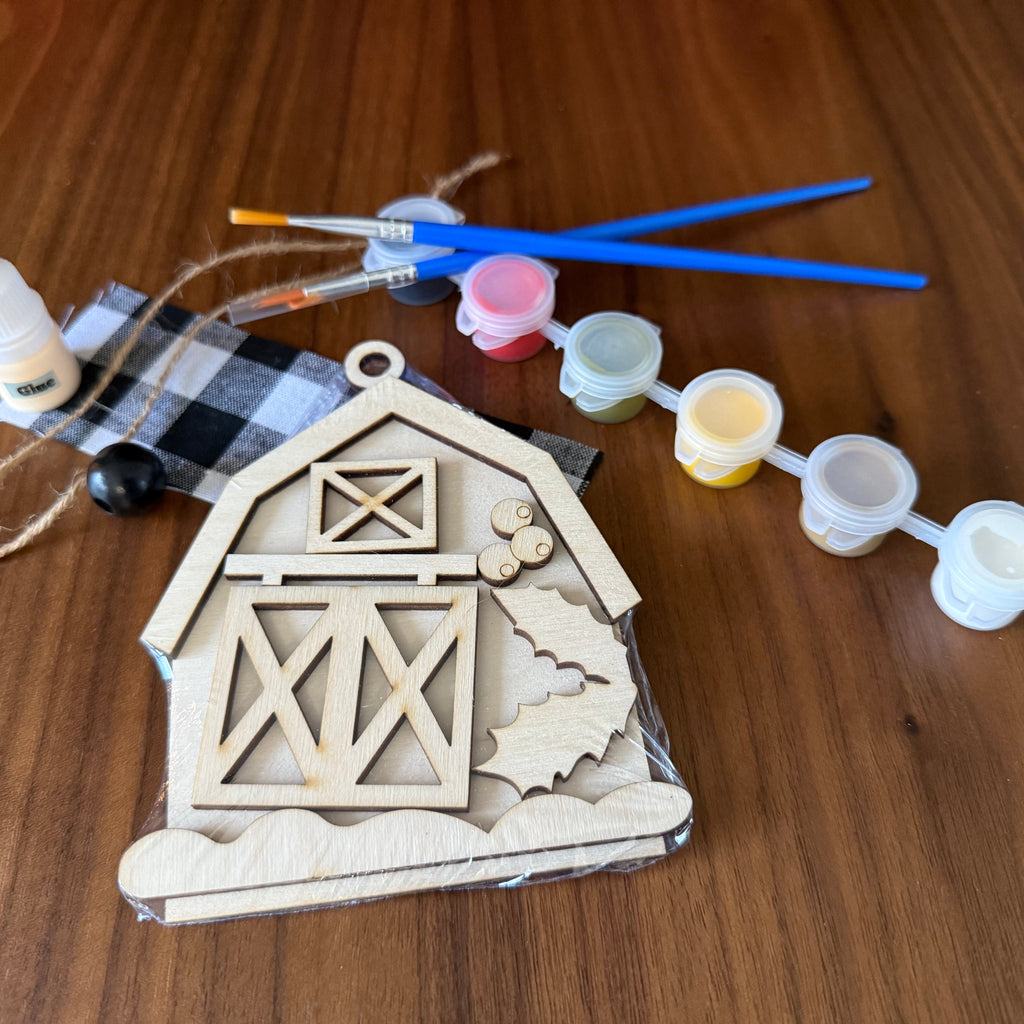 A wooden DIY craft kit for a barn, featuring an unfinished wooden barn door template, a black and white buffalo plaid fabric piece, twine, a bottle of wood glue, and five small paint pots with blue-handled paintbrushes, all arranged on a wooden tabletop.