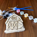 A wooden DIY craft kit for a barn, featuring an unfinished wooden barn door template, a black and white buffalo plaid fabric piece, twine, a bottle of wood glue, and five small paint pots with blue-handled paintbrushes, all arranged on a wooden tabletop.