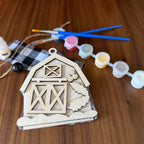 A wooden DIY craft kit for a barn, featuring an unfinished wooden barn door template, a black and white buffalo plaid fabric piece, twine, a bottle of wood glue, and five small paint pots with blue-handled paintbrushes, all arranged on a wooden tabletop.