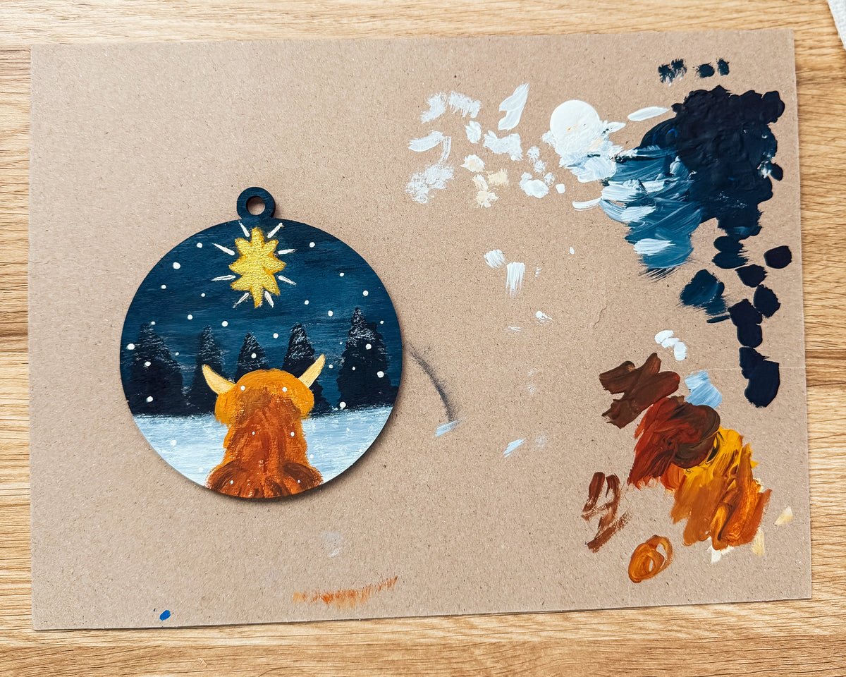 A circular painted ornament or pendant featuring a stylized orange animal in a snowy night scene, placed on a brown card over a wooden tabletop with paintbrushes and paint splatters visible around it.
