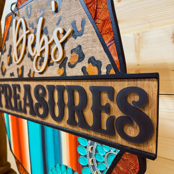 A decorative wooden sign with the words 'Debs TREASURES' in raised black letters. The sign has a rustic multicolor design with leopard print, turquoise stripes, and orange textured panels, mounted against a light wood plank wall.