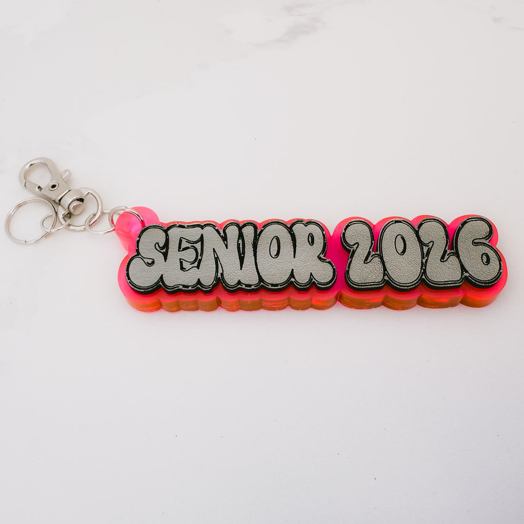 A 3D acrylic keychain featuring the text 'SENIOR 2026' in a gray bubble font with black outlines, layered over a vibrant pink and orange gradient base, complete with a silver lobster clasp on a white background.