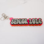 A 3D acrylic keychain featuring the text 'SENIOR 2026' in a gray bubble font with black outlines, layered over a vibrant pink and orange gradient base, complete with a silver lobster clasp on a white background.