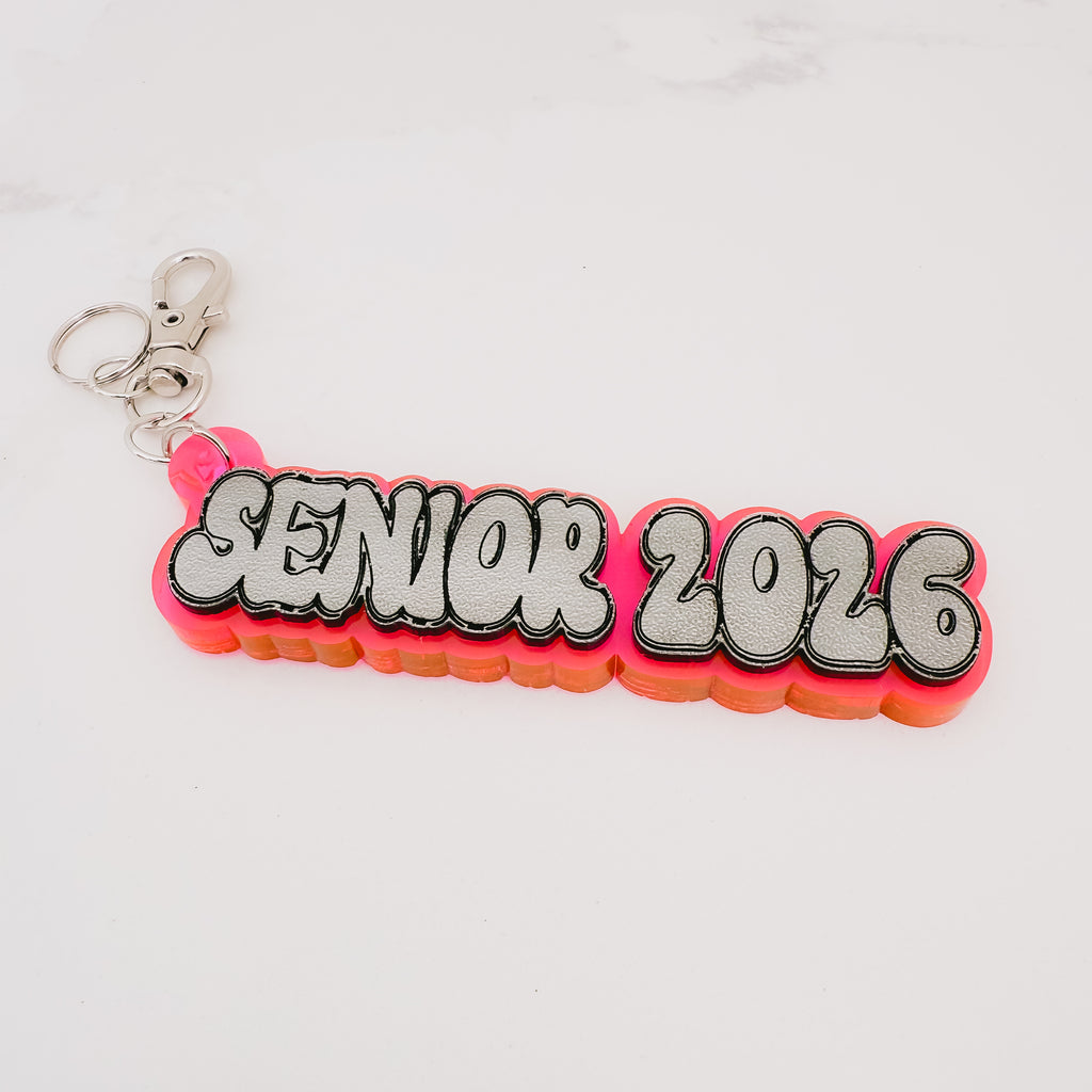 A custom 'SENIOR 2026' acrylic keychain featuring a bright pink and orange layered background with bold white letters outlined in black, attached to a silver split ring on a white marble surface.