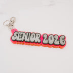 A custom 'SENIOR 2026' acrylic keychain featuring a bright pink and orange layered background with bold white letters outlined in black, attached to a silver split ring on a white marble surface.