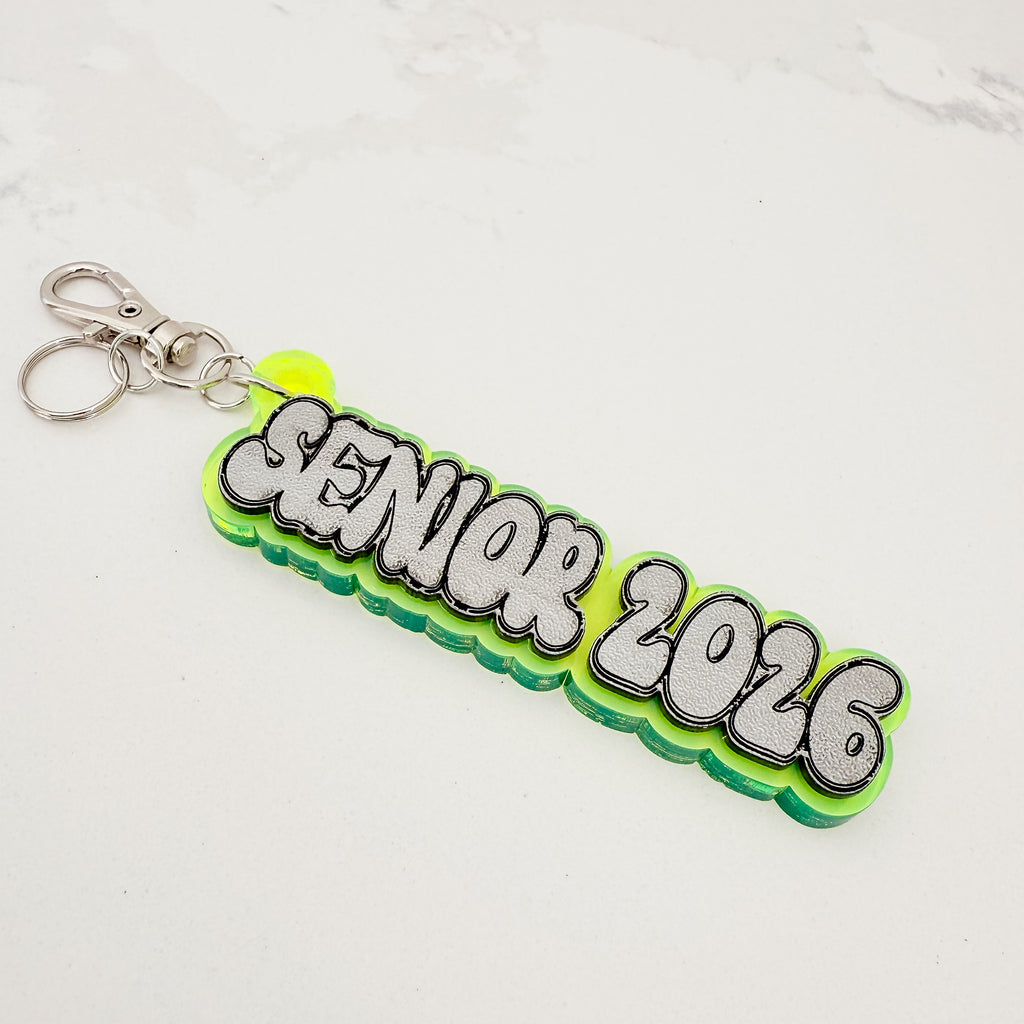 A custom 'SENIOR 2026' acrylic keychain with a bright green and neon yellow-green layered design, featuring 3D bubble letters with silver glitter fill and black outlines, attached to a silver split ring.