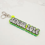 A custom 'SENIOR 2026' acrylic keychain with a bright green and neon yellow-green layered design, featuring 3D bubble letters with silver glitter fill and black outlines, attached to a silver split ring.