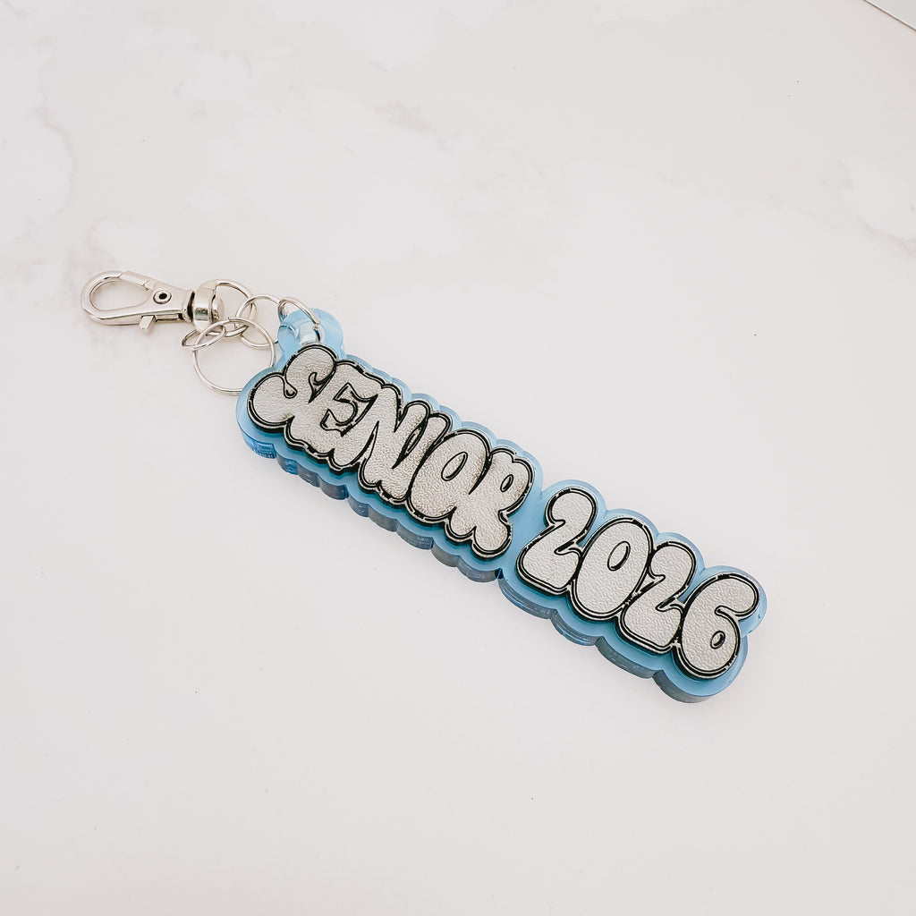 A custom 'SENIOR 2026' acrylic keychain with a light blue and white layered design, featuring a silver metal clasp on a white marble background.