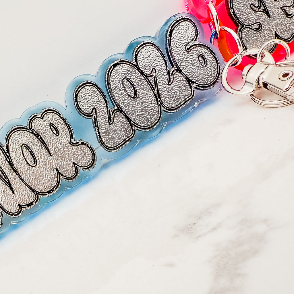 A close-up of a translucent blue acrylic keychain strap with textured silver lettering that reads '2026', attached to a silver metal clasp. Part of a pink keychain is visible in the background on a white marble surface.