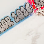 A close-up of a translucent blue acrylic keychain strap with textured silver lettering that reads '2026', attached to a silver metal clasp. Part of a pink keychain is visible in the background on a white marble surface.